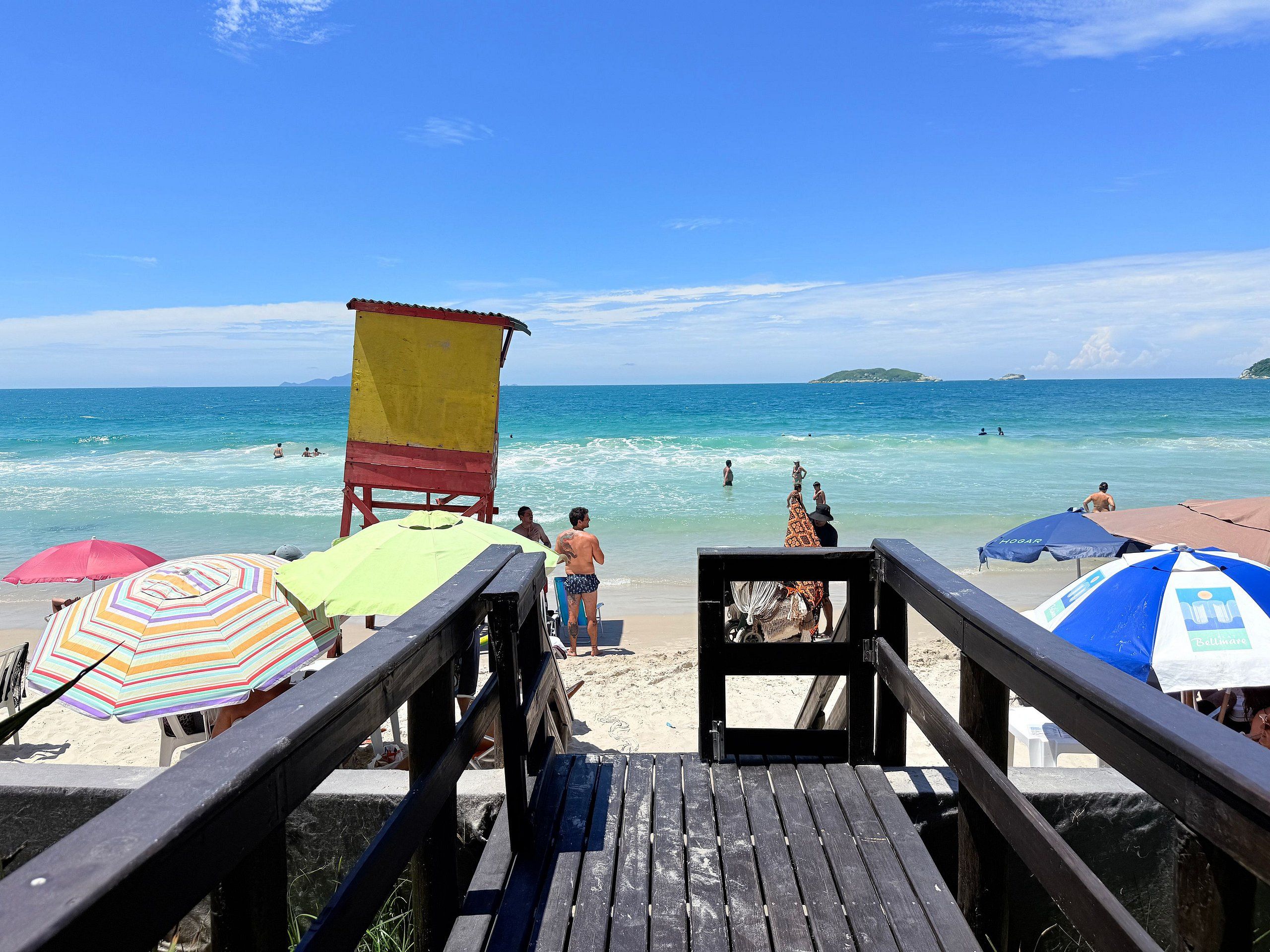 A wooden pier leading to a beach with umbrellas and a lifeguard tower.