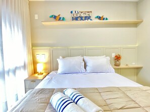 Habitación de hotel con cama, mesita de noche con lámpara, repisa con objetos decorativos y letrero en la pared.