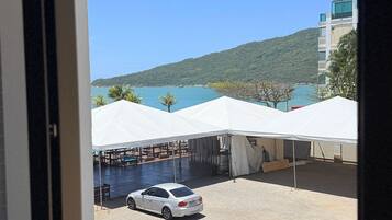 Visão pela janela de um hotel, mostrando um estacionamento, tendas brancas, um carro branco e um corpo d'água com uma colina ao fundo.