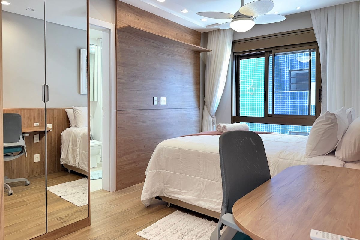 A hotel room with a bed, a desk, a chair, a ceiling fan, and a window with a city view.