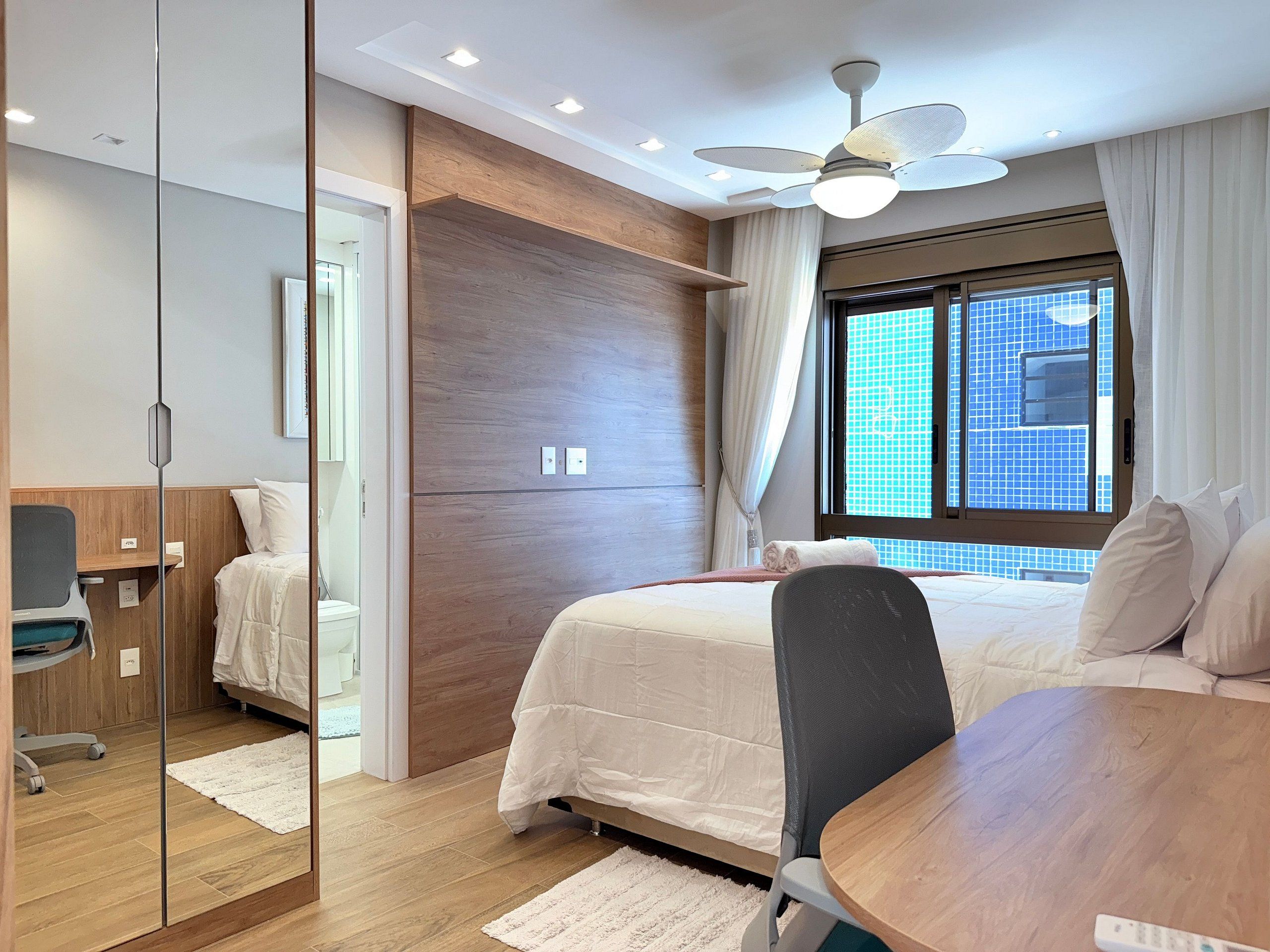 A hotel room with a bed, a desk, a chair, a ceiling fan, and a window with a city view.