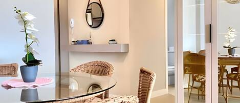 A dining area with a glass table, wicker chairs, a potted orchid, a mirror, and a view through a glass door.