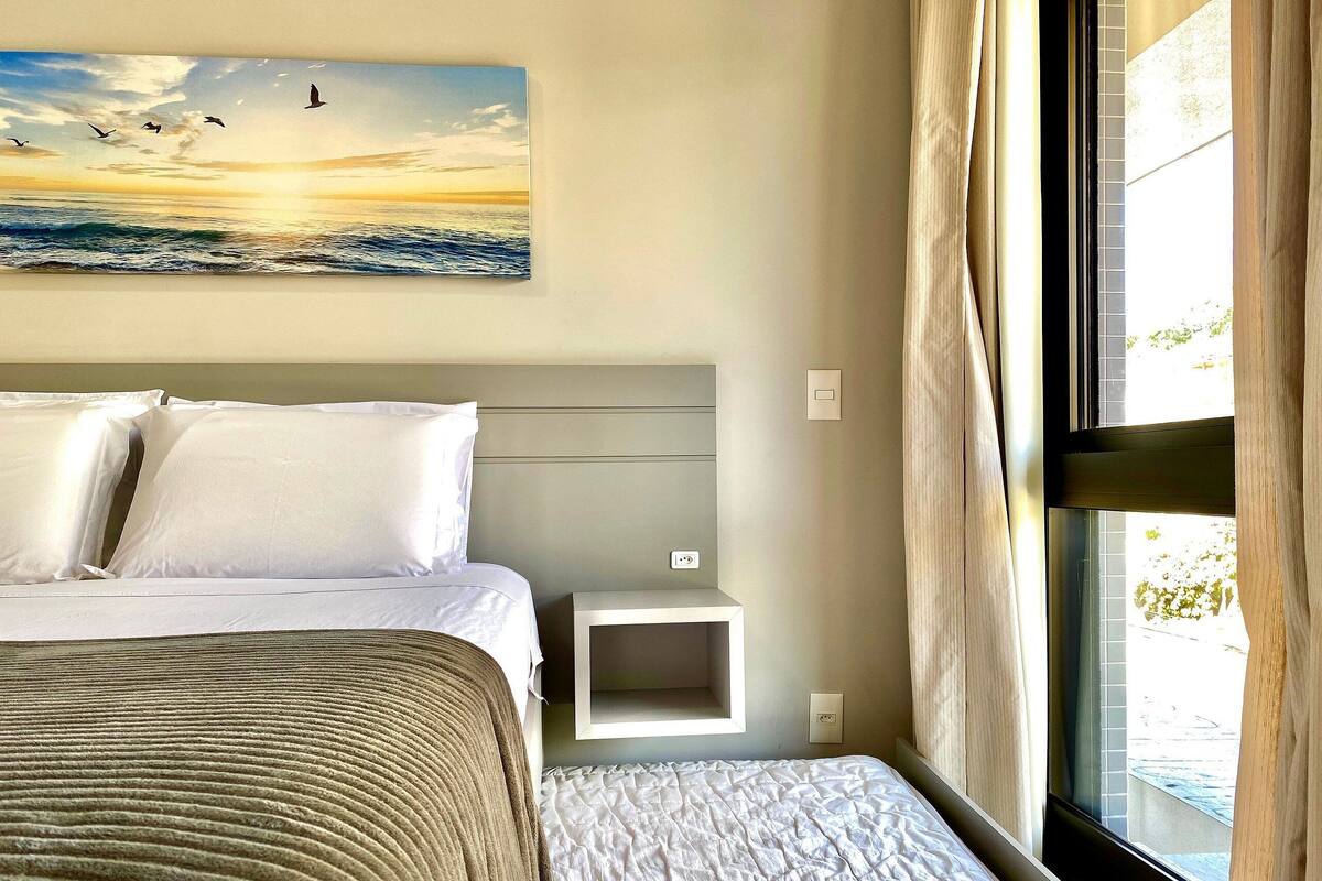 A hotel room with a bed, a bedside table, a nightstand, a painting of birds flying over water, and a window with curtains.