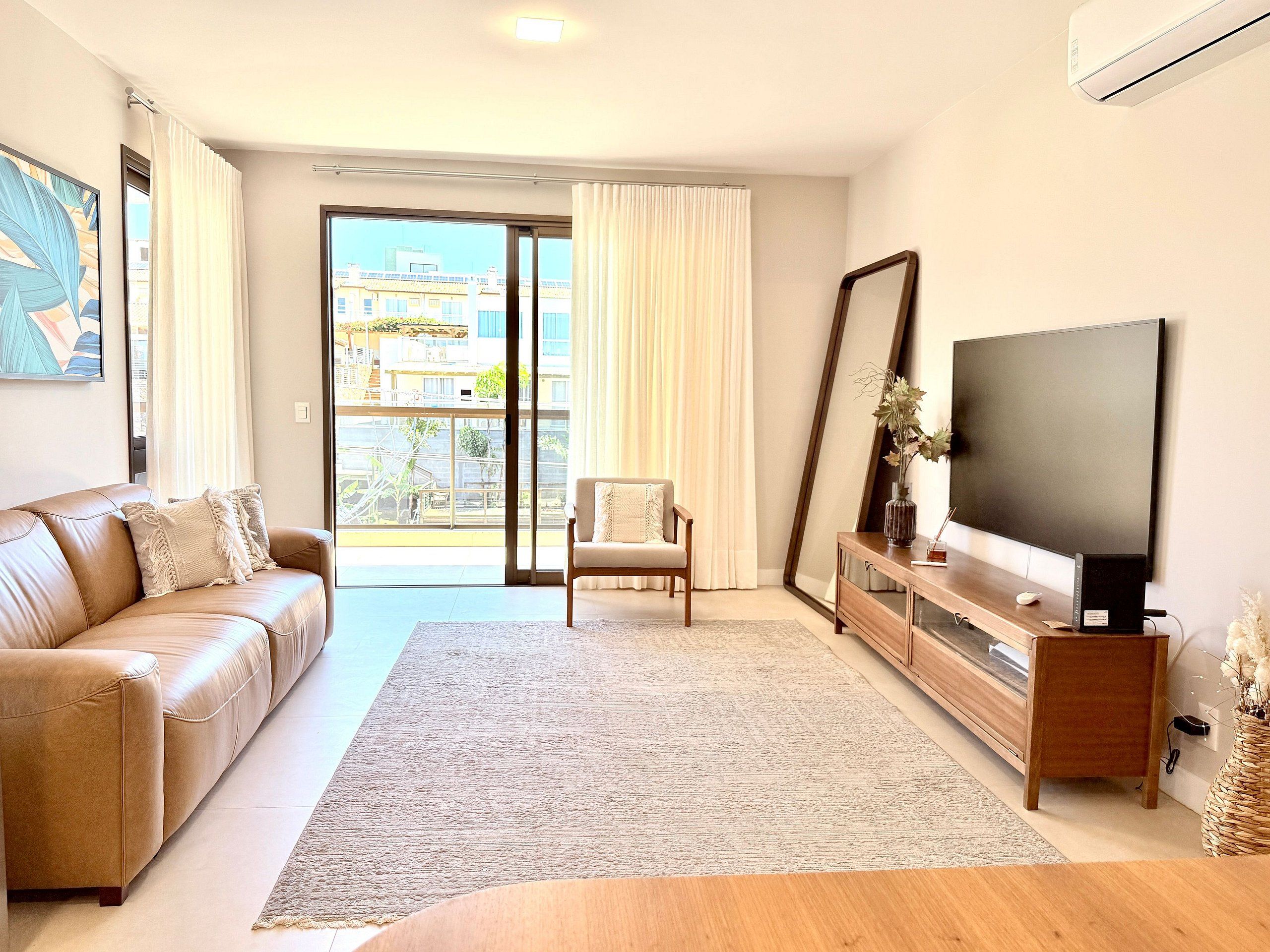 A modern living room with a leather sofa, a wooden coffee table, a flat-screen TV, and a large window with curtains.