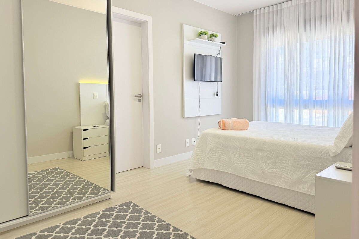A bedroom with a bed, a TV mounted on the wall, a bedside table, and a mirror.
