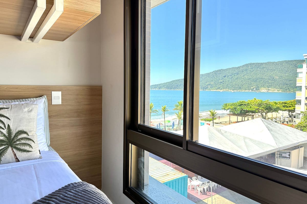 A bedroom with a large window, a bed with pillows, and a view of a beach and buildings.