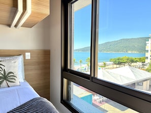 A bedroom with a large window, a bed with pillows, and a view of a beach and buildings. - Res Belvedere Beach Garden (Florianópolis)