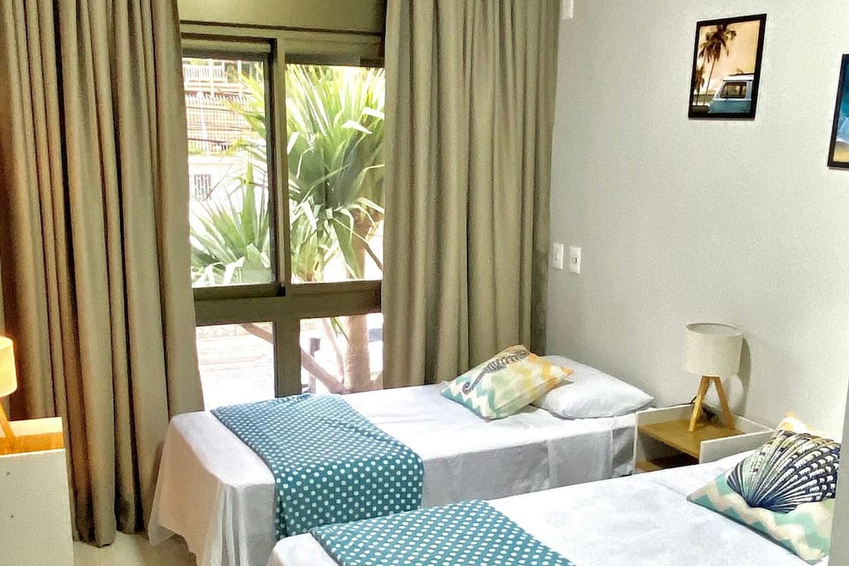 A hotel room with two beds, a desk, a chair, a window with curtains, and a small table with a lamp.