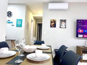 A dining area with a table set for four, a wall-mounted TV displaying a TV interface, and two framed artworks above the TV. - Res Belvedere Beach Garden (Florianópolis)