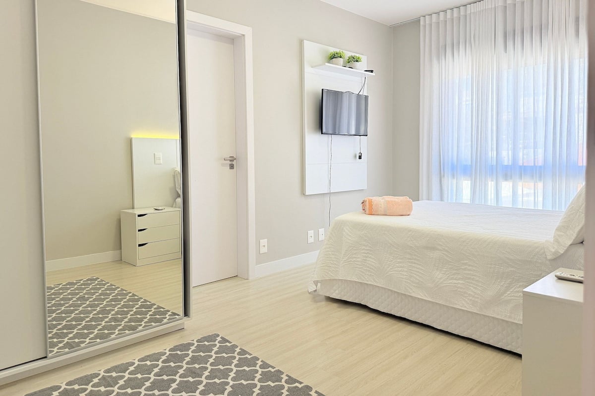 A bedroom with a bed, a TV mounted on the wall, a bedside table, and a mirror.