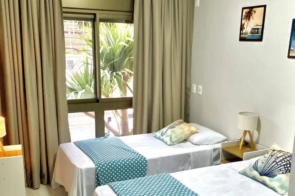 A hotel room with two beds, a desk, a chair, a window with curtains, and a small table with a lamp.
