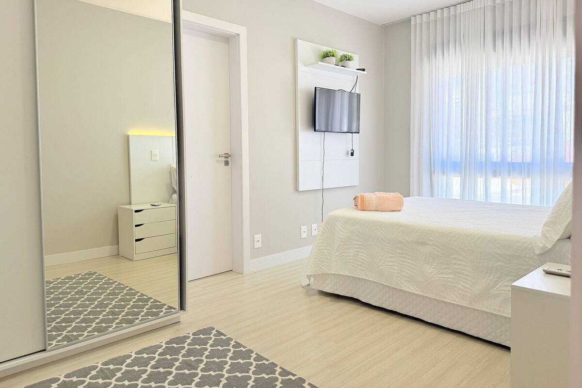 A bedroom with a bed, a TV mounted on the wall, a bedside table, and a mirror.