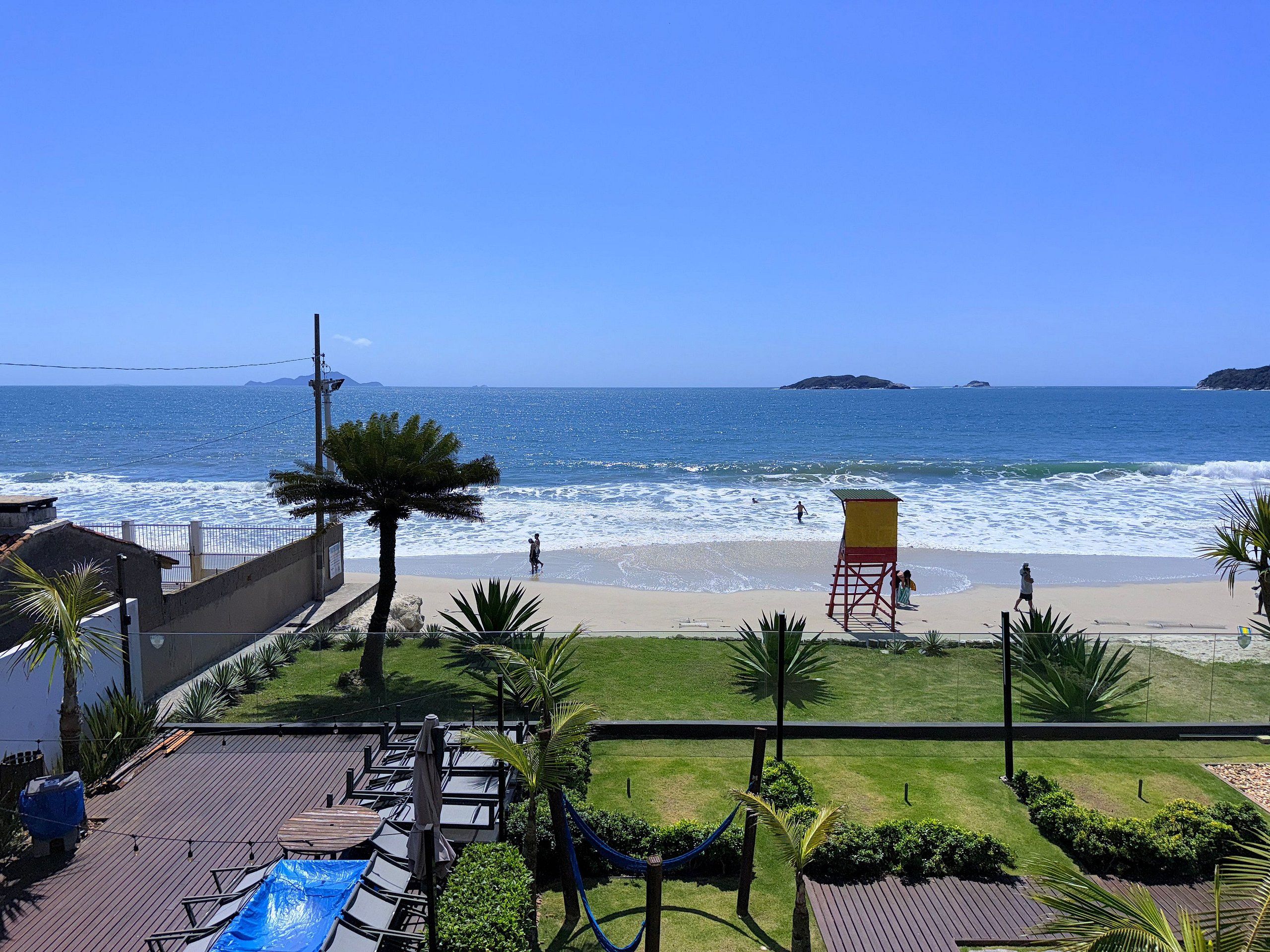 Vista de una playa con una torre de salvavidas, palmeras y un deck con sillas y sombrillas.