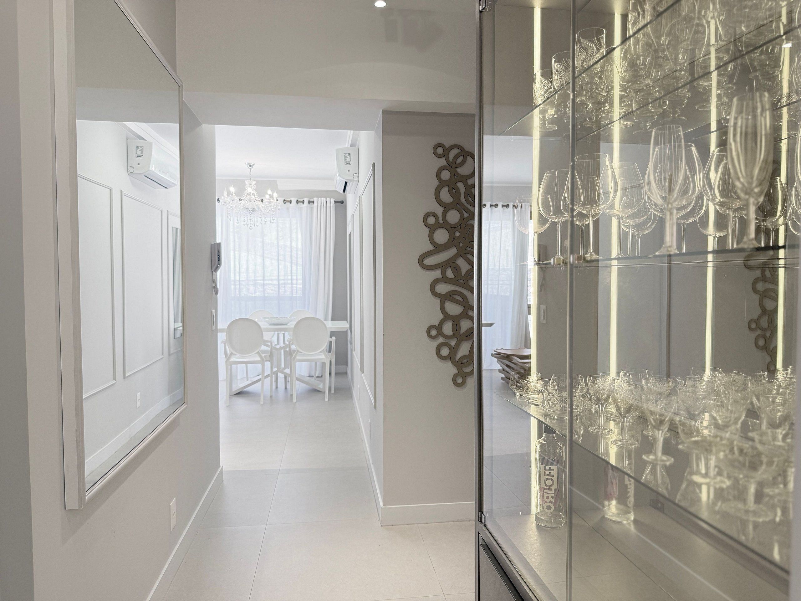 A corridor with a glass display case of wine glasses, a dining area with a chandelier, and a decorative wall piece.