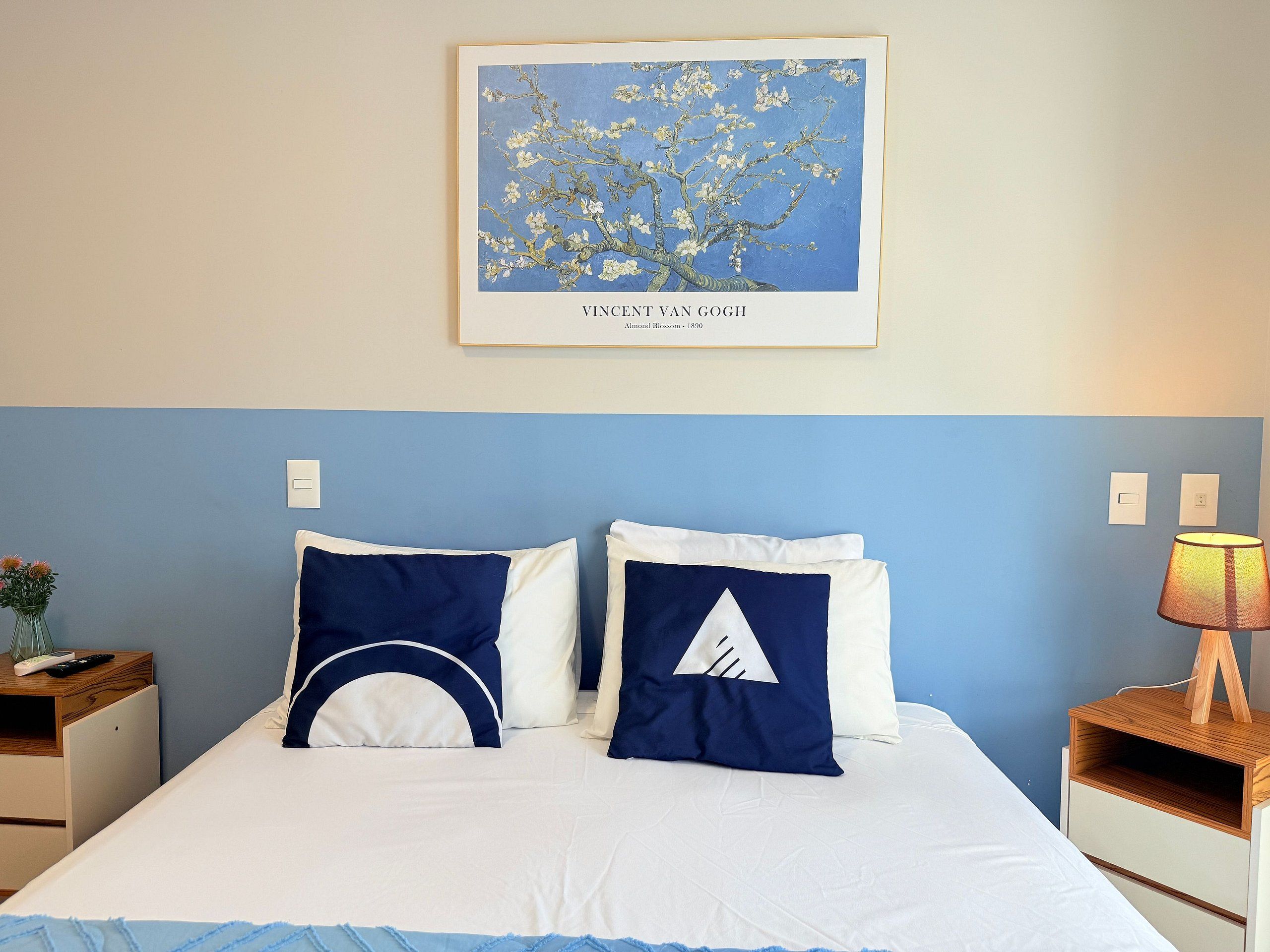A neatly made bed with white and navy blue pillows, a framed artwork above it, and a bedside table with a lamp.
