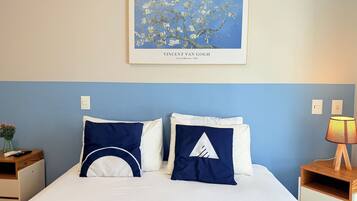 Una cama bien hecha con almohadas blancas y azul marino, una obra de arte enmarcada encima y una mesita de noche con una lámpara.