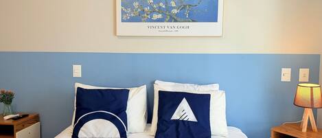A neatly made bed with white and navy blue pillows, a framed artwork above it, and a bedside table with a lamp.