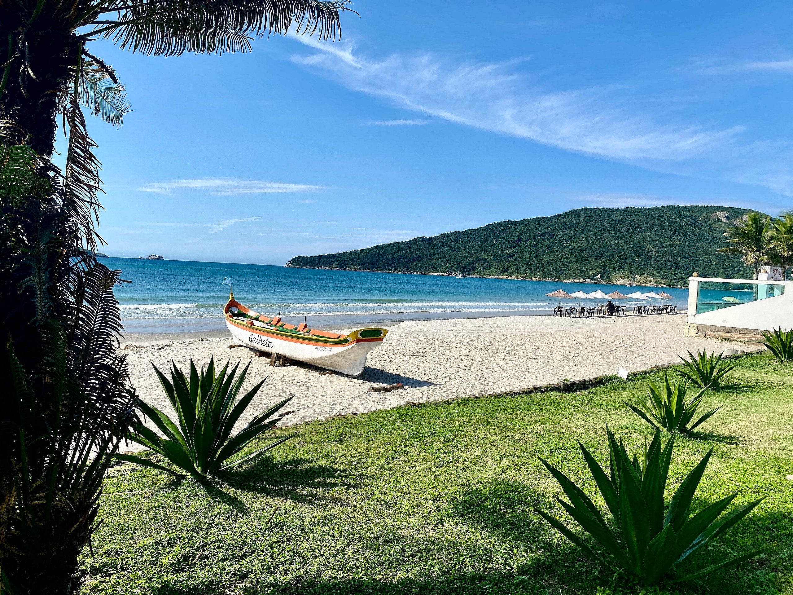 A boat named 'Galheta' on a beach with a grassy area, umbrellas, and a mountain in the background.