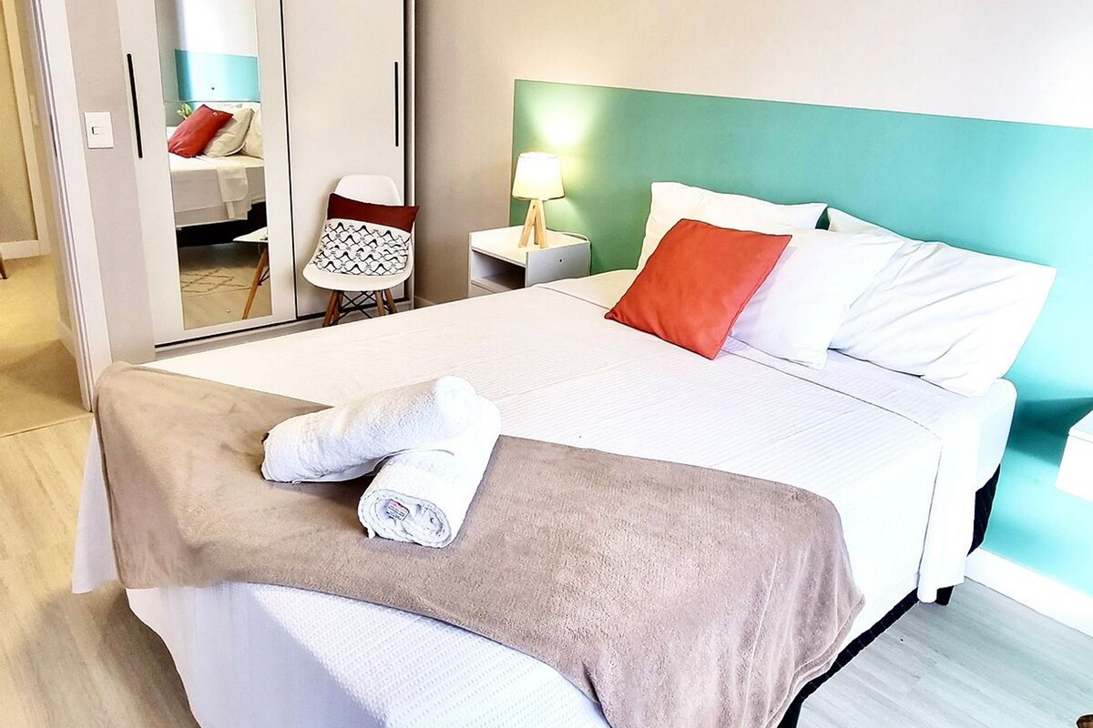 A neatly made bed with a white and brown blanket, a red pillow, and a white towel. There is a nightstand with a lamp, a chair, and a wardrobe with a mirror. The room has light wooden flooring and a green accent wall.