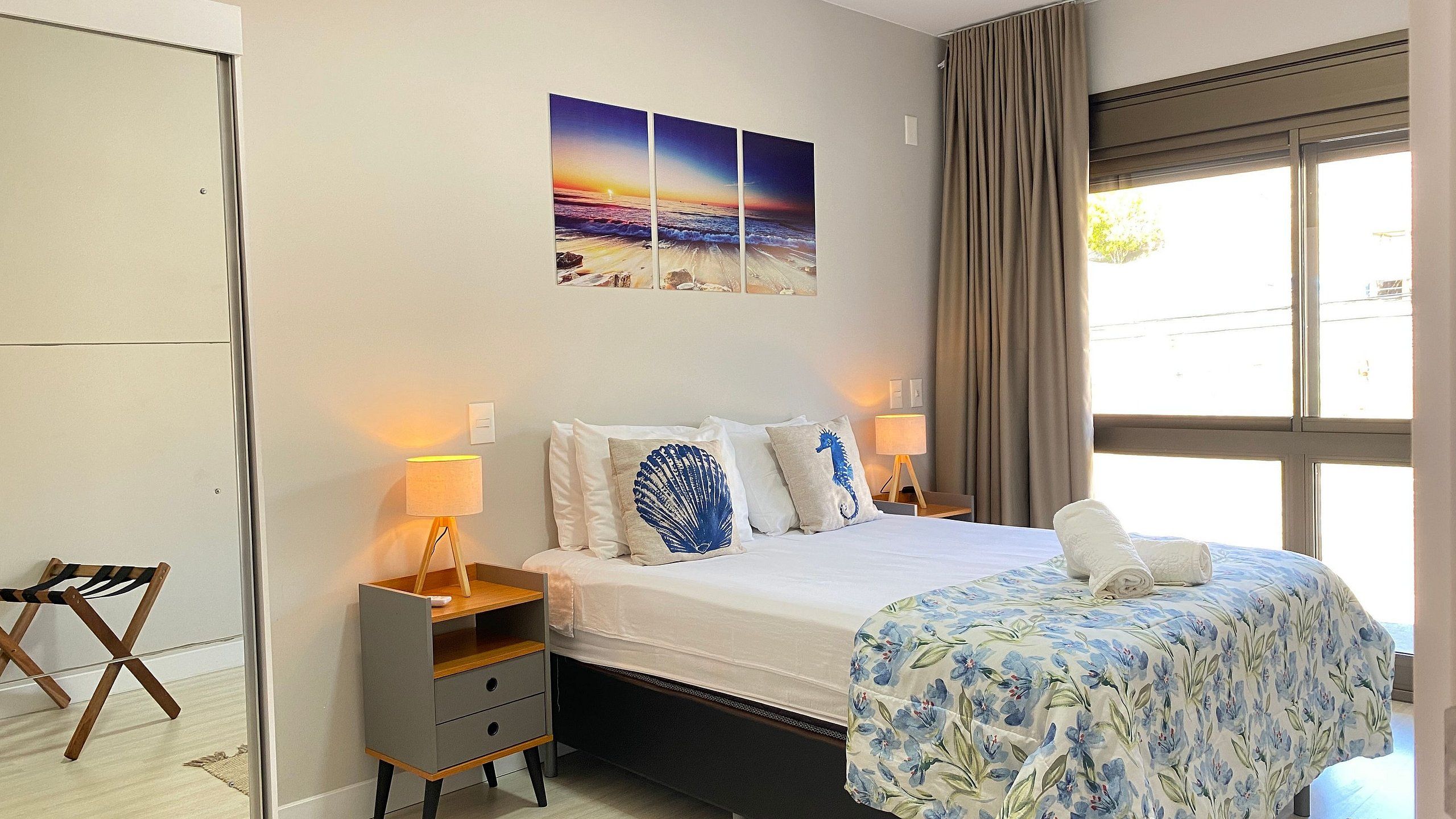 Une chambre d’hôtel comprenant un lit, des lampes de chevet, une table de chevet, une chaise, une fenêtre avec des rideaux et une œuvre d’art murale sur le thème de la plage.