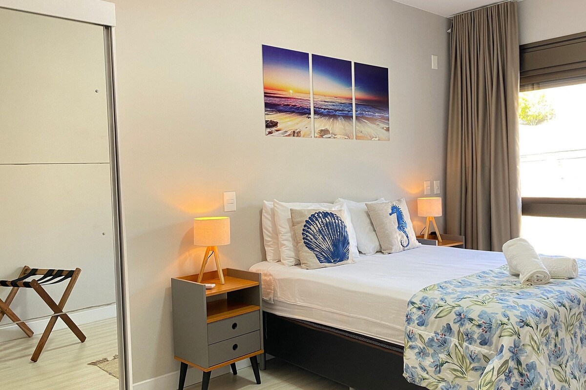 Habitación de hotel con cama, lámparas de noche, mesita de noche, una silla, ventana con cortinas y una obra de arte con temática playera.