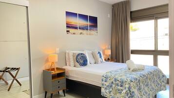 Une chambre d’hôtel comprenant un lit, des lampes de chevet, une table de chevet, une chaise, une fenêtre avec des rideaux et une œuvre d’art murale sur le thème de la plage.