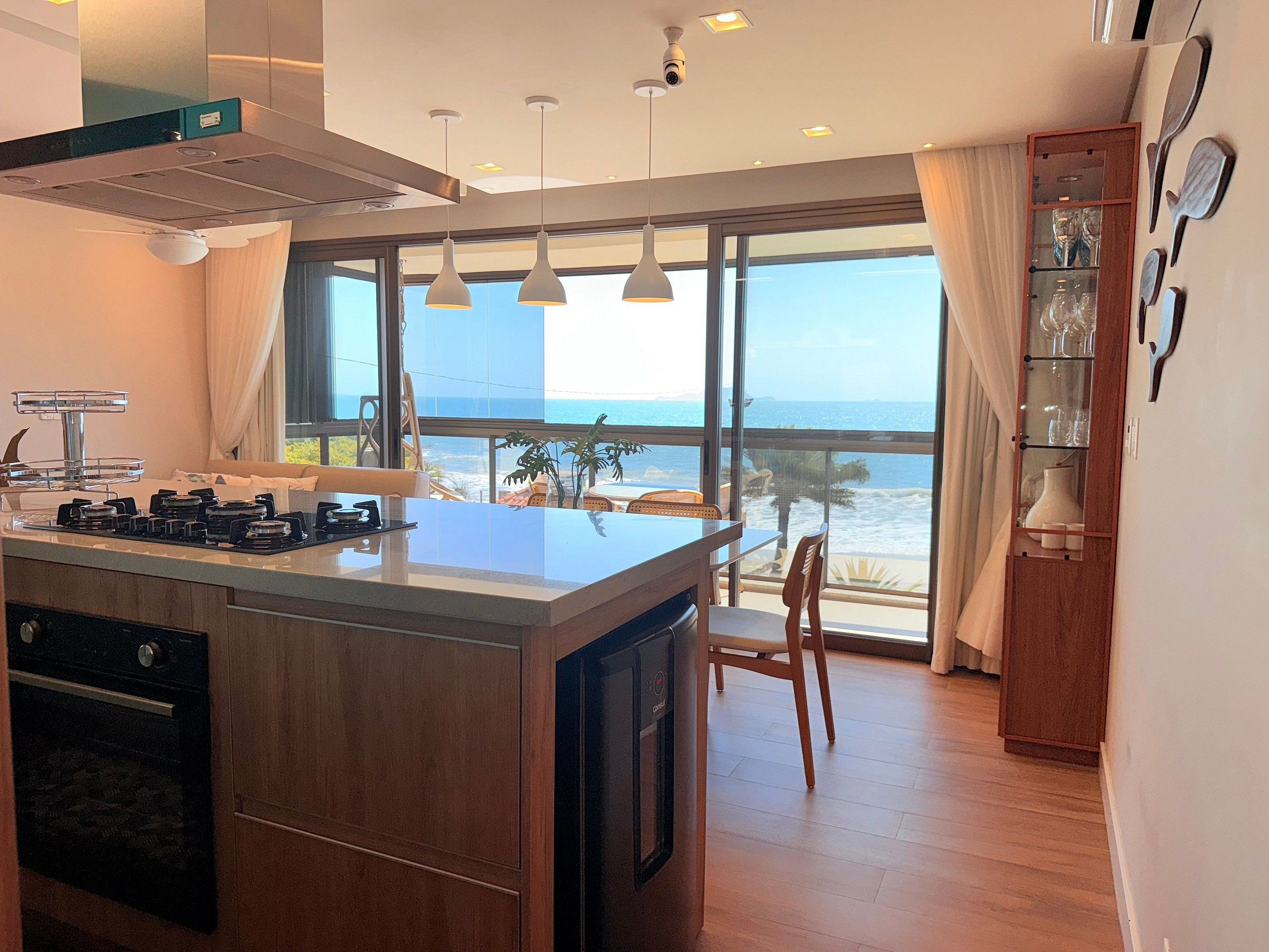 A modern kitchen with a central island, a dining area with a view of the ocean, and a wooden cabinet with glassware.