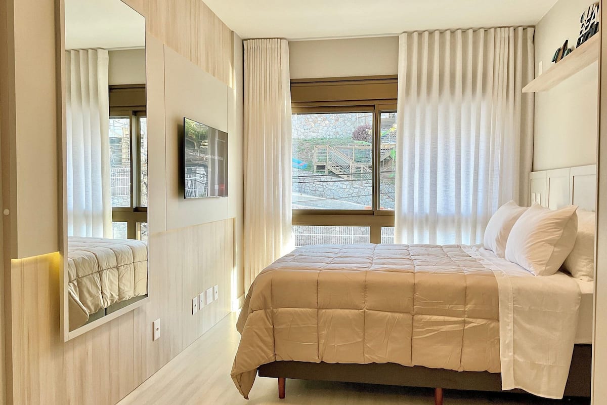 A bedroom with a bed, a TV, a window with curtains, and a view of the outside.