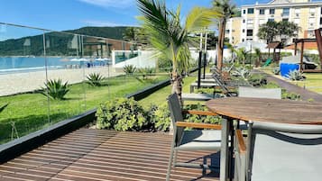 Een houten terras met een tafel en stoelen, uitkijkend op een strand en een gebouw met veel ramen.