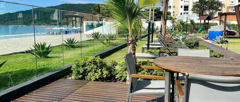 Una terraza de madera con una mesa y sillas, con vista a una playa y un edificio de múltiples ventanas.