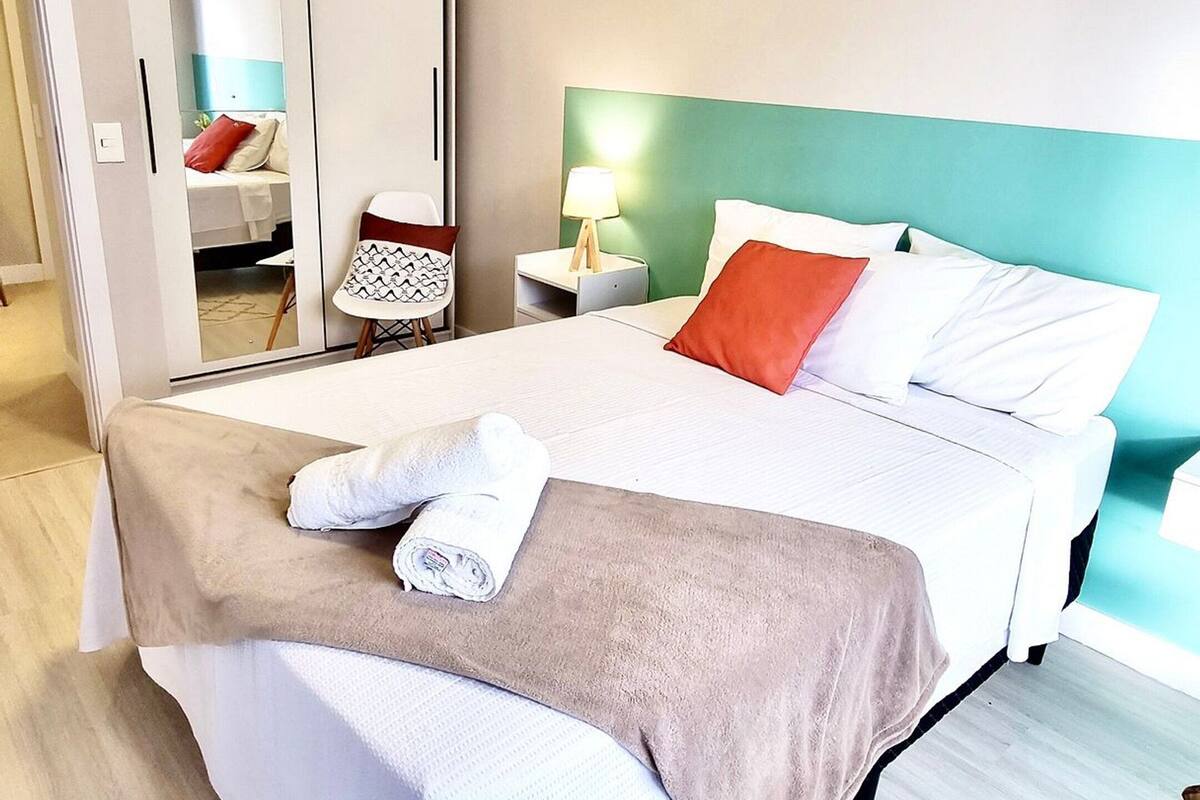 A neatly made bed with a white and brown blanket, a red pillow, and a white towel. There is a nightstand with a lamp, a chair, and a wardrobe with a mirror. The room has light wooden flooring and a green accent wall.