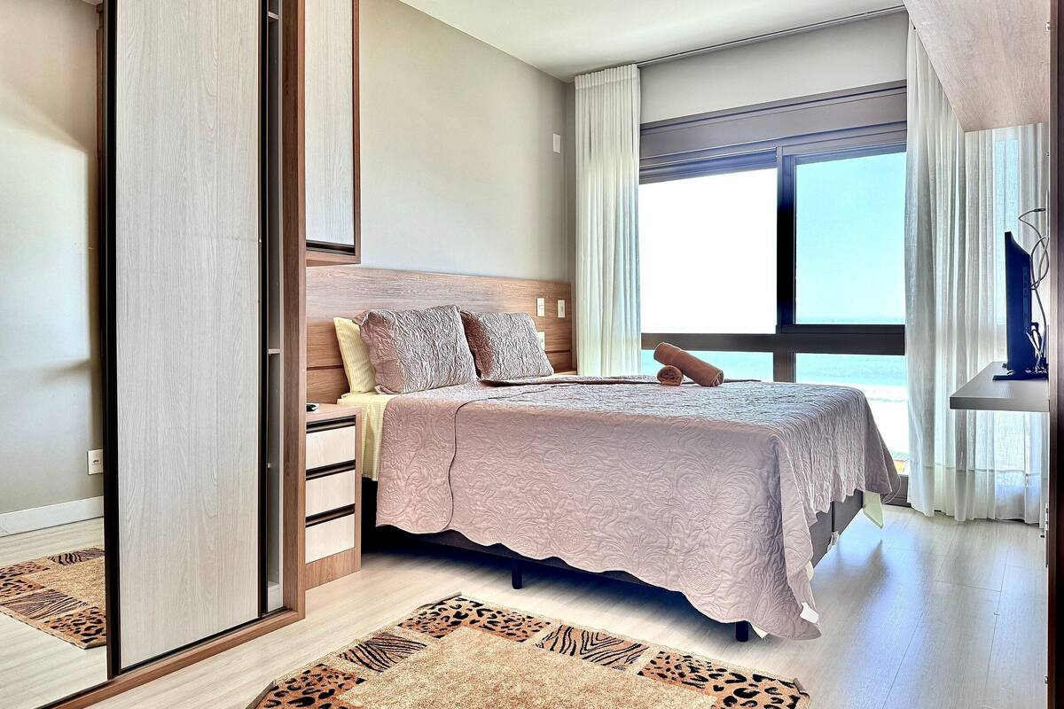 A bedroom with a bed, a wardrobe, a window with curtains, and a view of the sea.