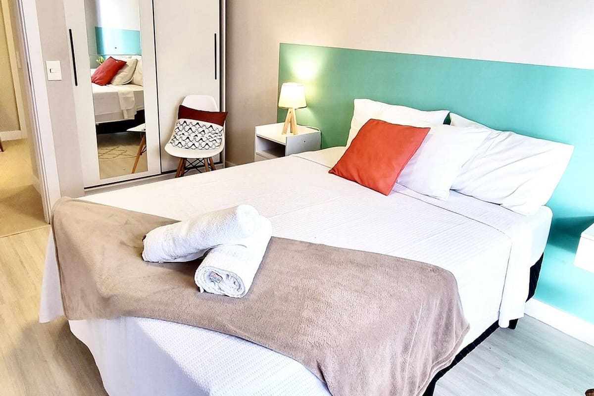 A neatly made bed with a white and brown blanket, a red pillow, and a white towel. There is a nightstand with a lamp, a chair, and a wardrobe with a mirror. The room has light wooden flooring and a green accent wall.