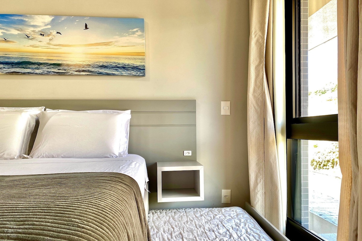 A hotel room with a bed, a bedside table, a nightstand, a painting of birds flying over water, and a window with curtains.
