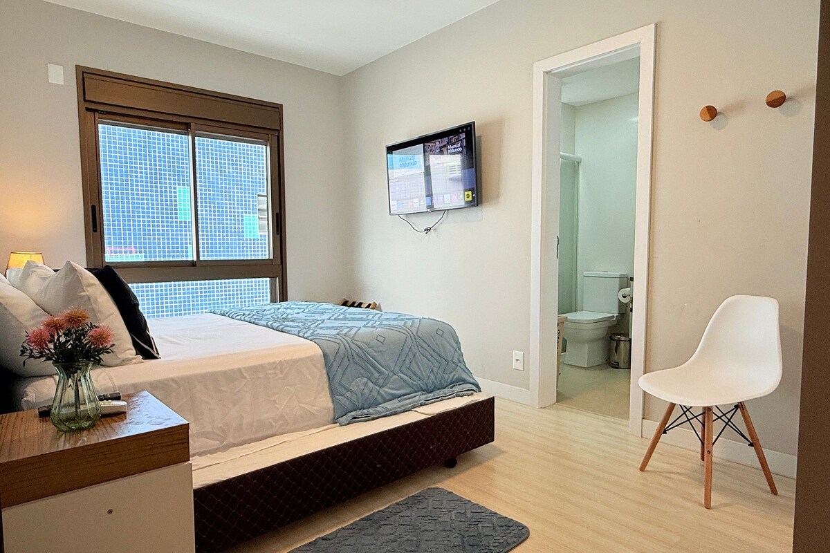 Une chambre à coucher comprenant un lit, un bureau, une chaise, un téléviseur, et une salle de bain visible à travers une porte ouverte.
