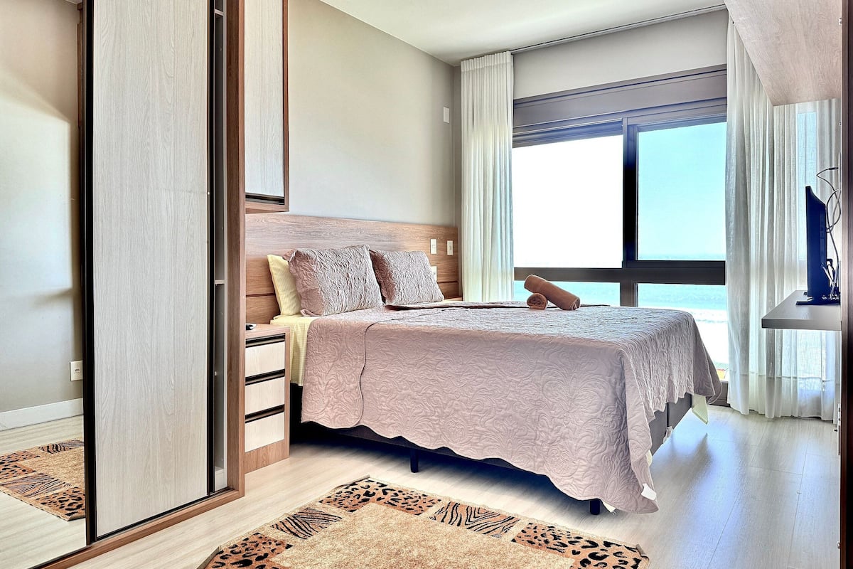 A bedroom with a bed, a wardrobe, a window with curtains, and a view of the sea.