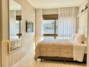 A bedroom with a bed, a TV, a window with curtains, and a view of the outside.