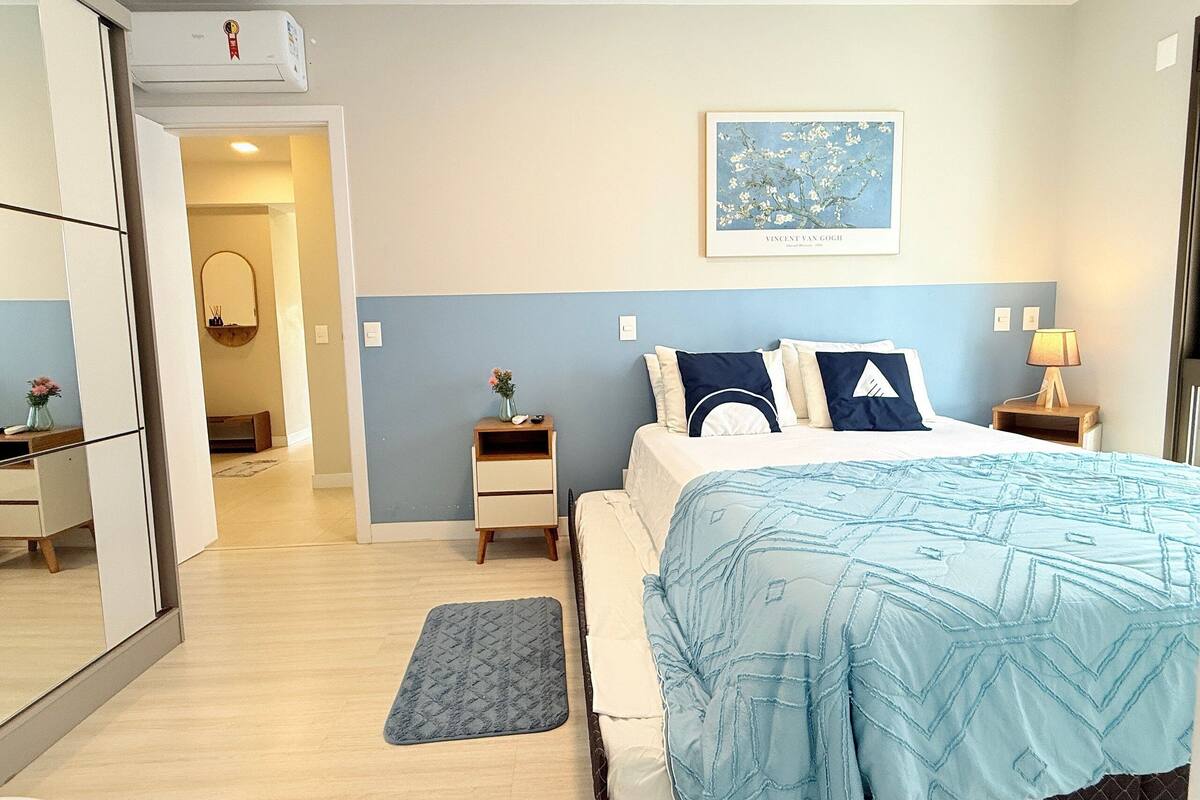 A bedroom with a bed, bedside tables, a nightstand, a wall-mounted air conditioner, a framed picture, and a door leading to another room.