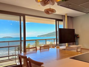 A modern room with a large window overlooking a beach and ocean, a dining area with a table and chairs, and a flat-screen TV on a stand.