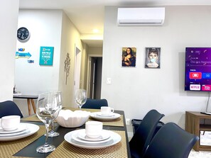 Un comedor con una mesa dispuesta para cuatro, un televisor montado en la pared mostrando una interfaz, y dos obras de arte enmarcadas encima del televisor.