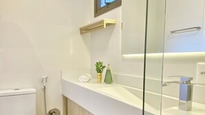 A modern bathroom with a toilet, a glass shower enclosure, a white sink with a mirror, and a small shelf with toiletries. - Res Belvedere Beach Garden (Florianópolis)