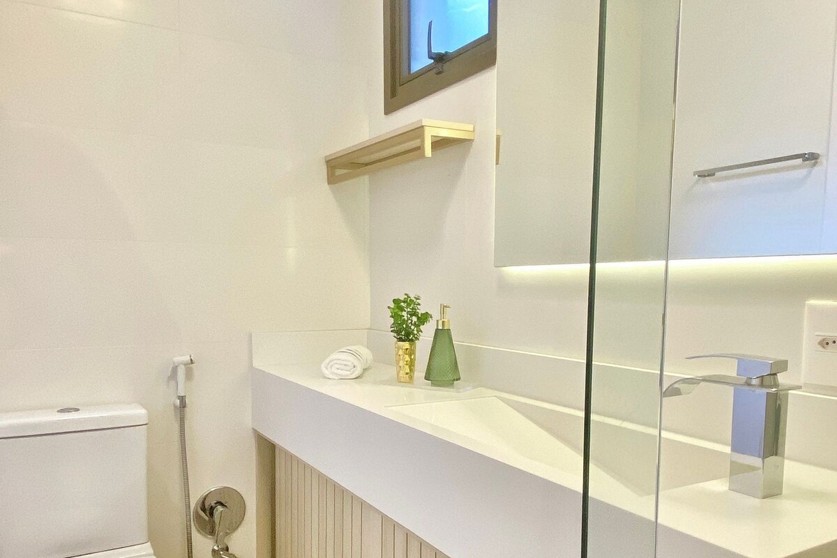 Um banheiro moderno com vaso sanitário, box de vidro, pia branca com espelho e prateleira pequena com produtos de higiene pessoal.