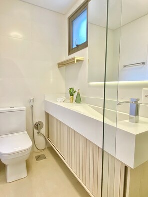 Um banheiro moderno com vaso sanitário, box de vidro, pia branca com espelho e prateleira pequena com produtos de higiene pessoal.