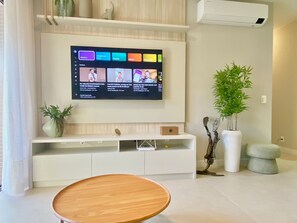 Een moderne woonkamer met een flatscreen televisie aan de muur, een houten salontafel, een witte tv-kast en twee potplanten.