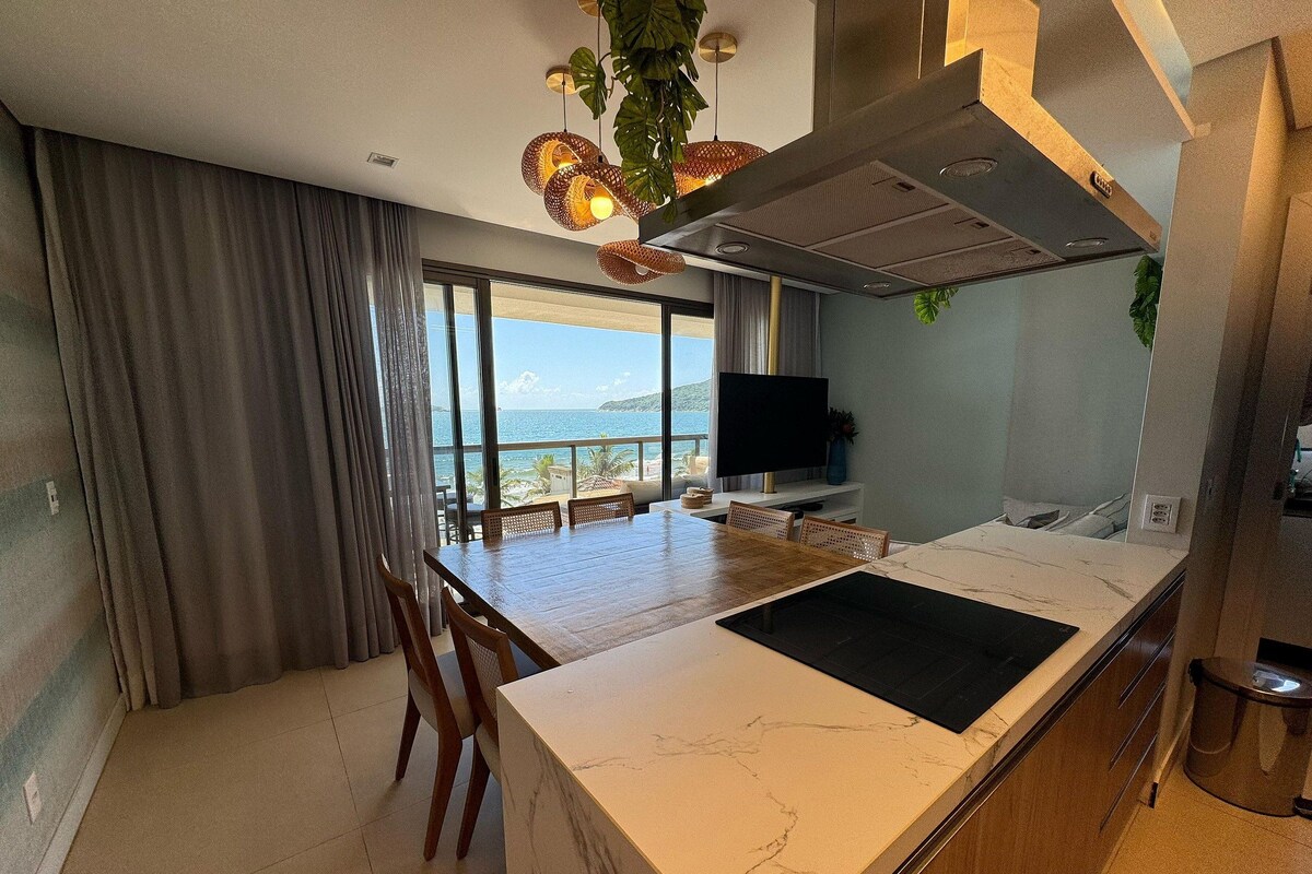 Ett modernt kök med en bänkskiva i marmor, en matplats med stolar, en tv och utsikt över havet.