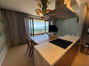 A modern kitchen with a marble countertop, a dining area with chairs, a TV, and a view of the sea. - Res Belvedere Beach Garden (Florianópolis)