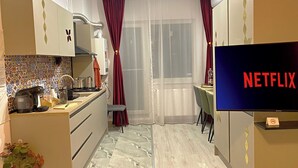Una cucina compatta con zona pranzo, una TV che mostra il logo di Netflix e una finestra con tende leggere.