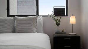 Un lit bien fait, agrémenté d’oreillers à rayures, une table de chevet avec une lampe, un vase avec une plante et une fenêtre donnant sur un paysage urbain.