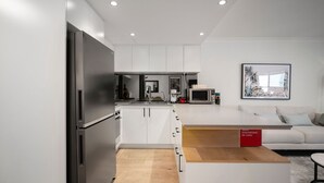 Une cuisine moderne avec des meubles blancs, un réfrigérateur en acier inoxydable, un four à micro-ondes et une machine à café. On y trouve une table à manger en bois et un canapé, avec un panneau rouge en arrière-plan.