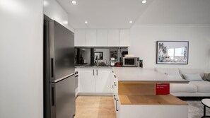 Eine moderne Küche mit weißen Schränken, einem Edelstahlkühlschrank, einer Mikrowelle und einer Kaffeemaschine. Ein Holztisch und eine Couch befinden sich im Raum, im Hintergrund ist ein rotes Schild zu sehen.